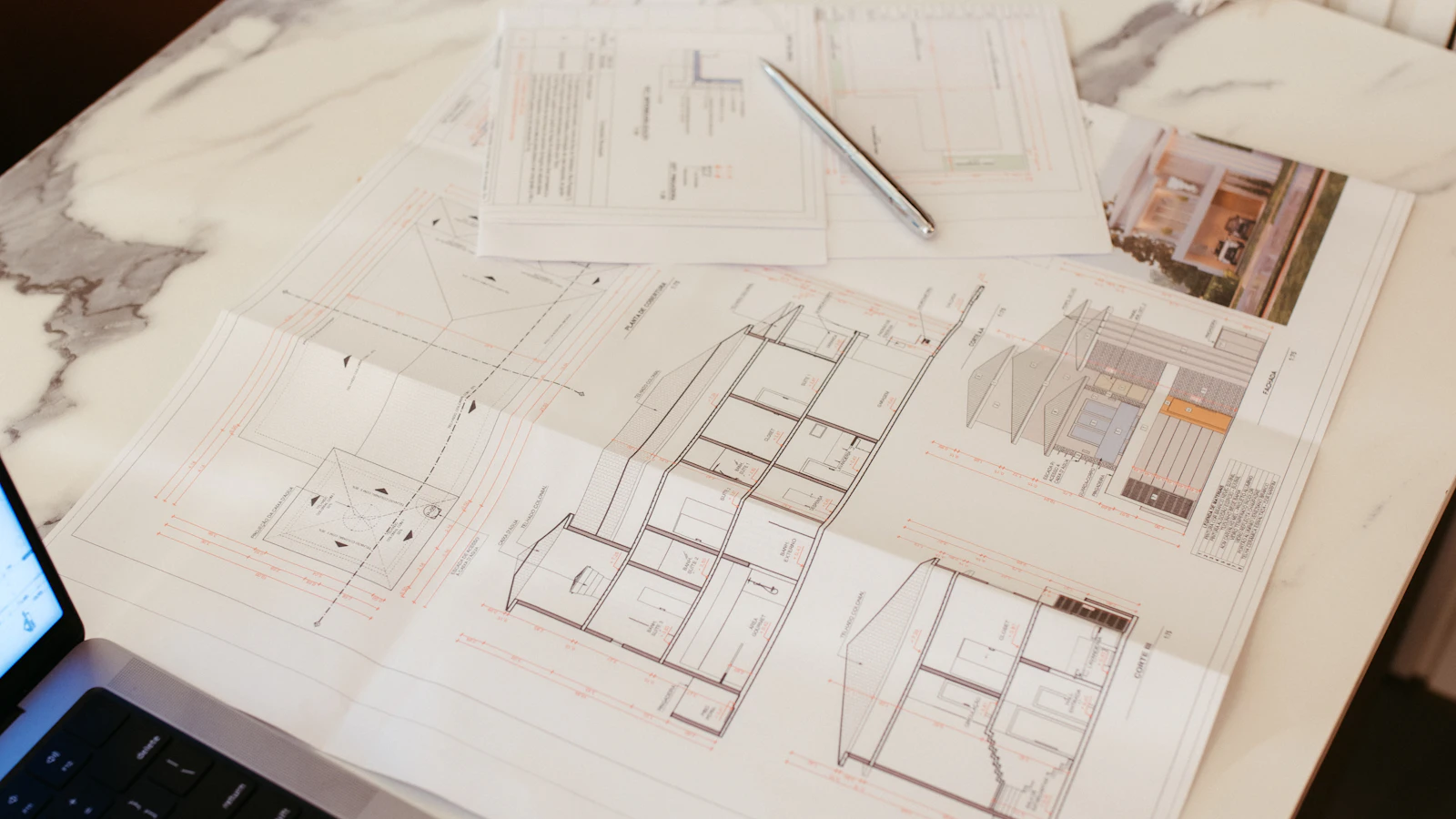 Architectural plans, a laptop and a pen arranged on a worktable.
